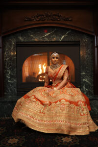 Bride in ornate orange lehenga by fireplace.